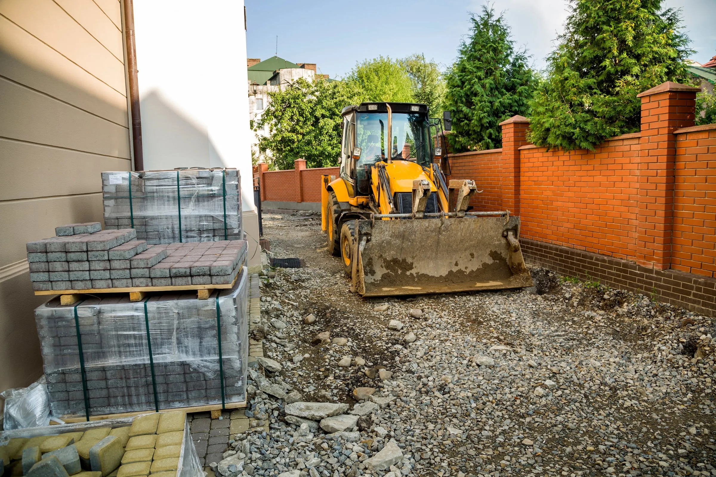 A yellow backhoe loader on a prepared gravel base beside stacked plastic-wrapped interlock pavers, ready for driveway installation.