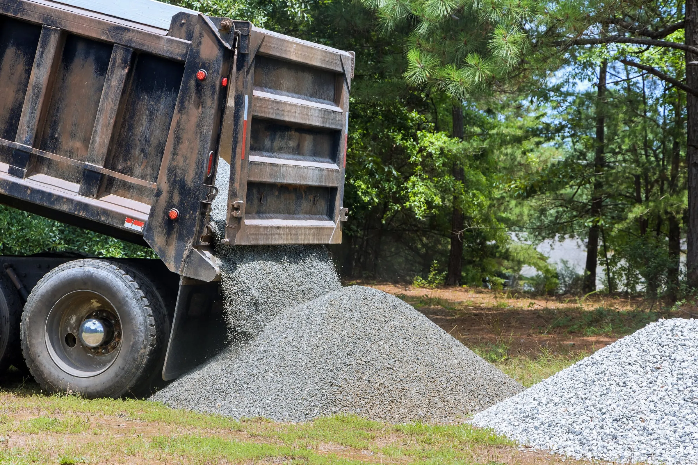 A dump truck unloading a large pile of gravel in a wooded area.