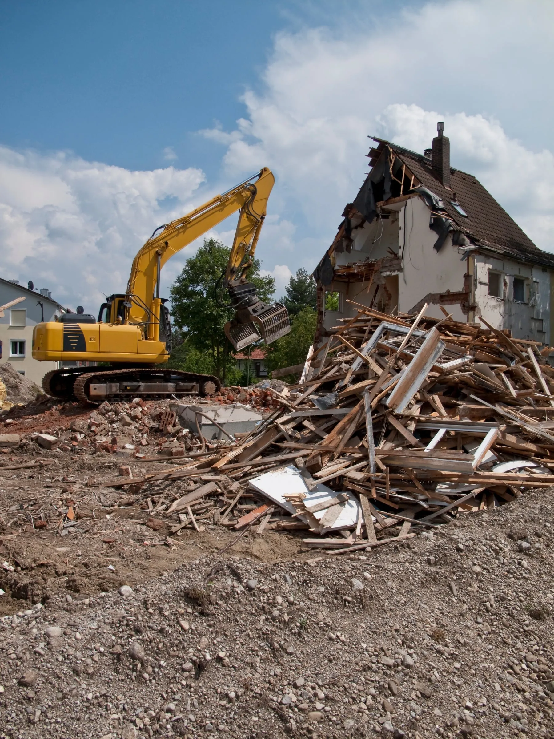 A yellow excavator with a grapple attachment tears down a residential building, with splintered wood framing piled beside the partially demolished structure.