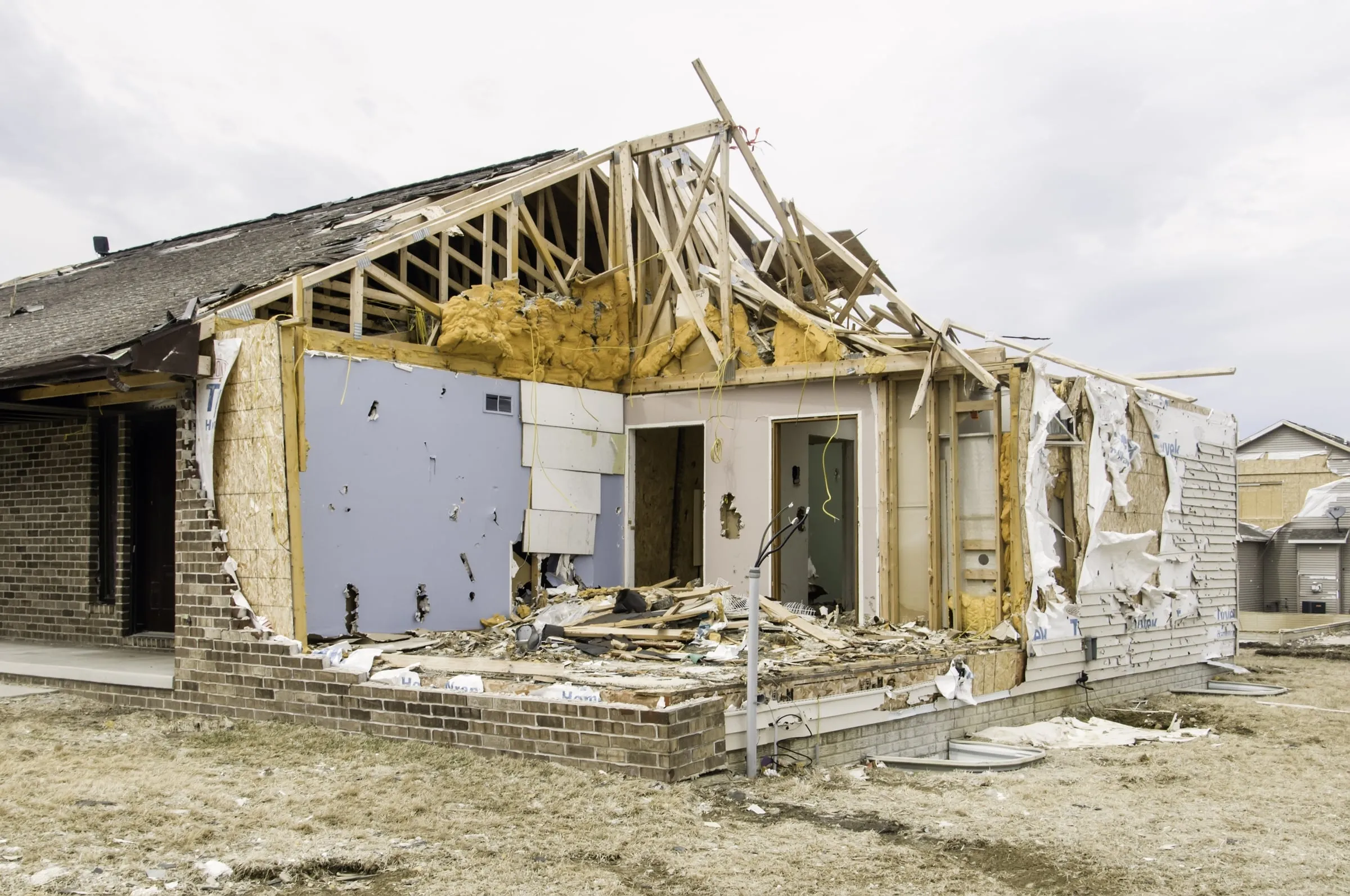 A residential home stripped to its exposed wood wall framing after severe storm damage, with torn insulation and debris scattered across the yard.