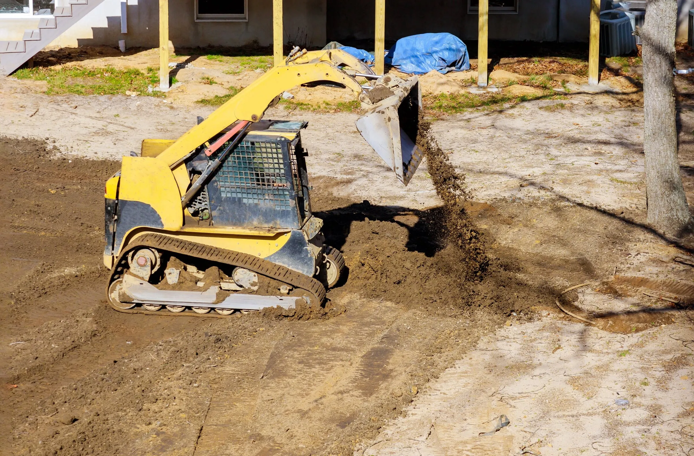 A yellow skid steer loader dumping a bucket of dirt near a residential house.